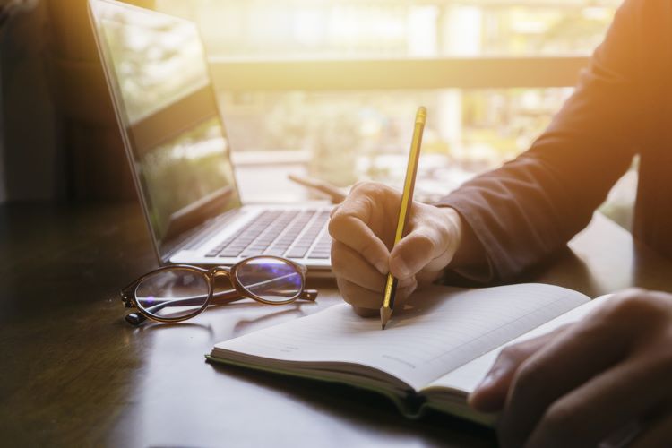 An online student writes in a notebook while referencing a laptop.